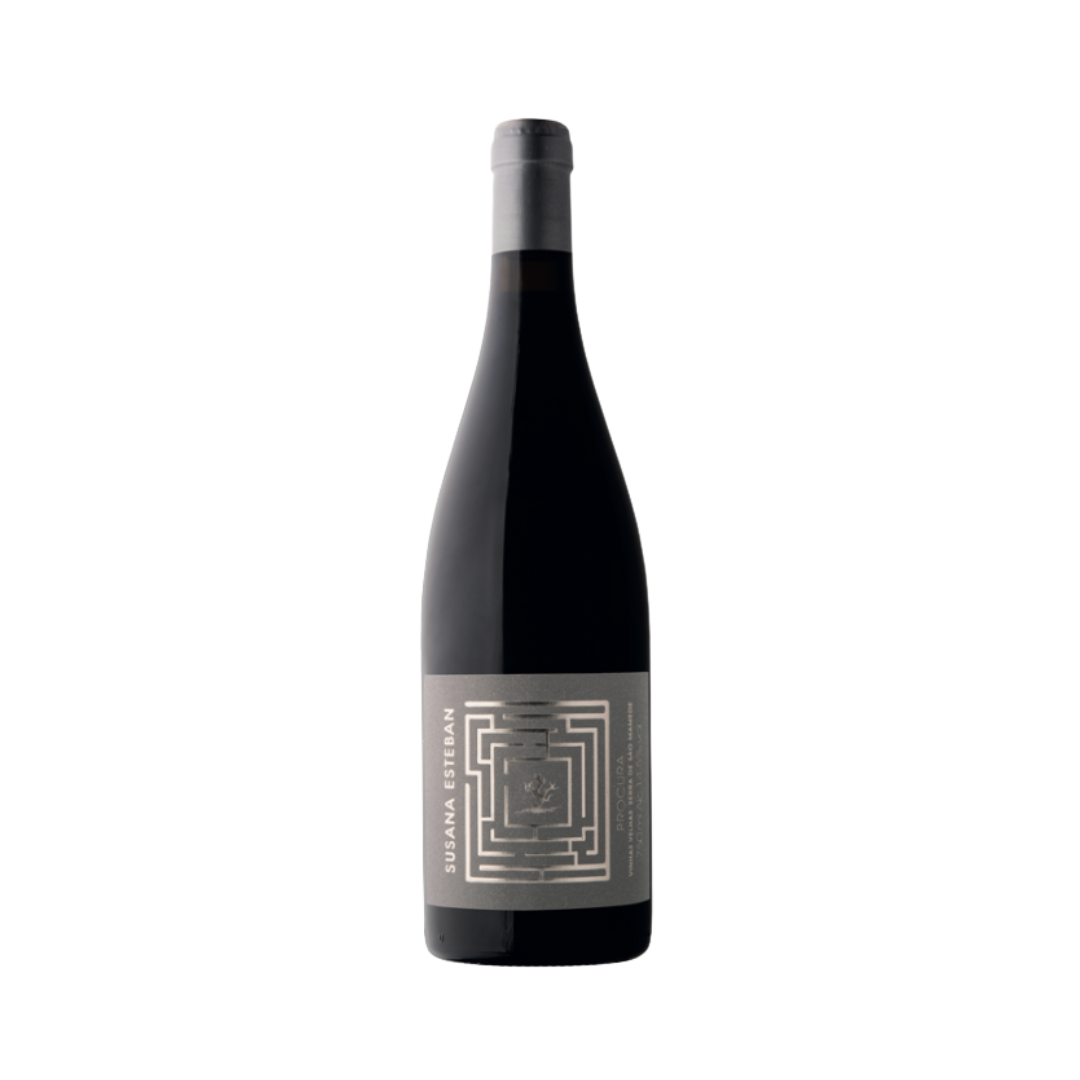 Bottle of Susana Esteban Procura Tinto 2019 on a dark wood table with mountain herbs and black fruits in the background, evoking the wine's powerful structure and origin from old vines in Serra de São Mamede.